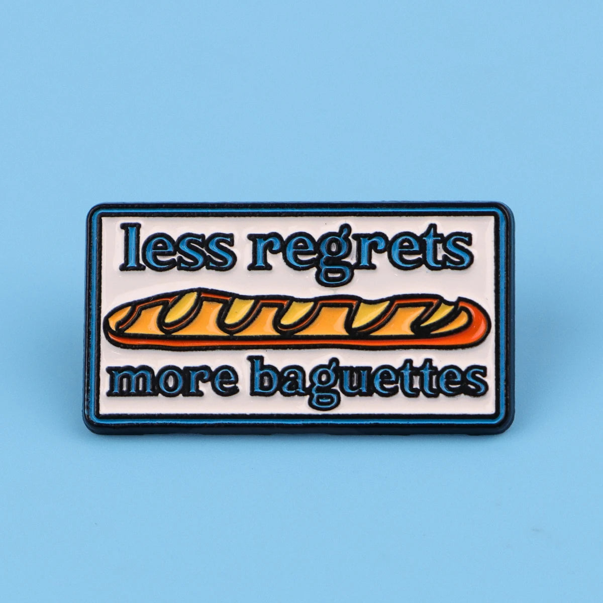 An enamel pin featuring the text less regrets more baguettes on a rectangular background.