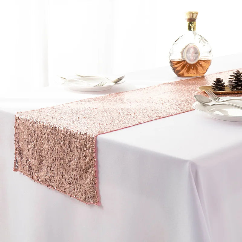 A rose gold sequin table runner displayed on a white tablecloth with a bottle of amber liquid and silverware.