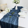 A deep blue semi-sheer gauze table runner draped elegantly on a white tablecloth with white plates and floral arrangements.