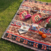 A woven blanket with geometric patterns and fringe laid on green grass, featuring musical instruments and a pink object.
