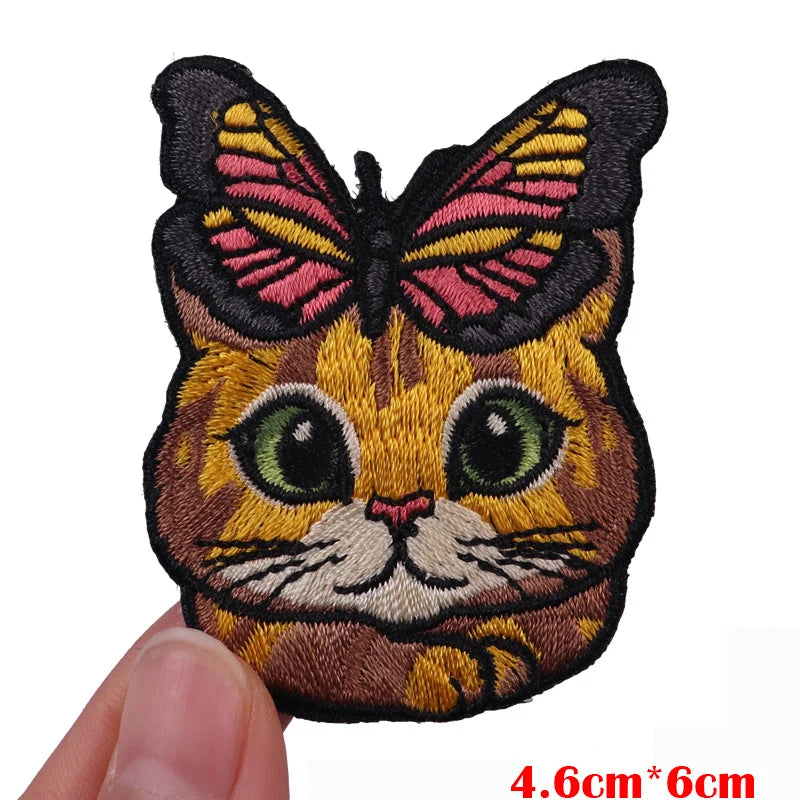 An embroidered patch of a cartoon cat face with pink and yellow wings.