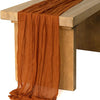 A rust orange rustic gauze table runner draped over a wooden table.