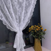 A sheer white floral lace curtain panel draped over a glass surface next to a vase of yellow flowers.