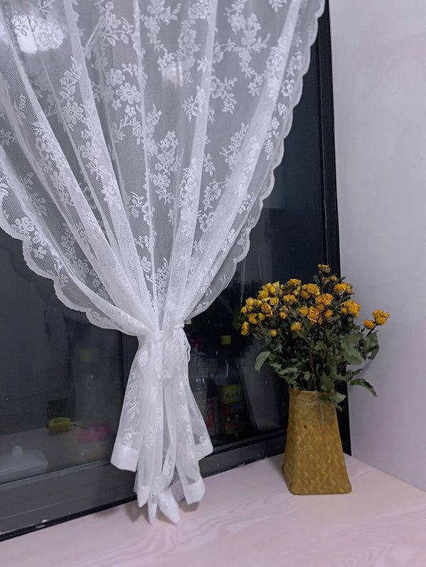 A sheer white floral lace curtain panel draped over a glass surface next to a vase of yellow flowers.