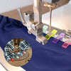 Close-up of colorful plastic sewing clips being used on blue fabric by a sewing machine needle.
