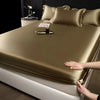 A close-up of a satin fitted sheet in gold color covering a mattress, with pillows and a bottle on top.