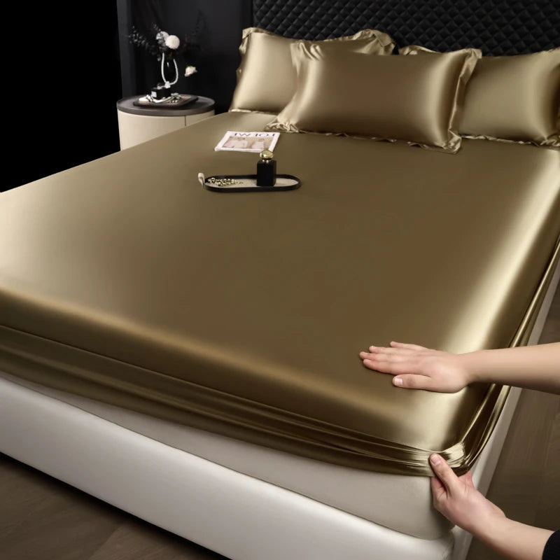 A close-up of a satin fitted sheet in gold color covering a mattress, with pillows and a bottle on top.