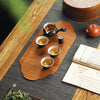 A set of ceramic tea cups and a small bamboo tea mat are arranged on a dark gray surface.