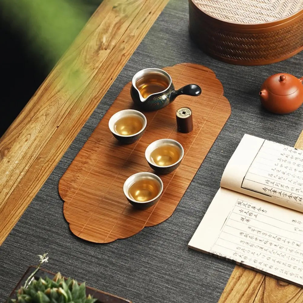 A set of ceramic tea cups and a small bamboo tea mat are arranged on a dark gray surface.