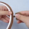 Hands are holding a round wooden embroidery hoop with a white inner ring and a gold-colored metal tool.