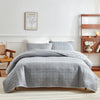 A gray quilted cotton bedspread set with matching pillows on a bed against a white wall.