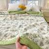 A hand holds a light green floral printed quilt with a yellow trim on a white bed, surrounded by soft pillows.