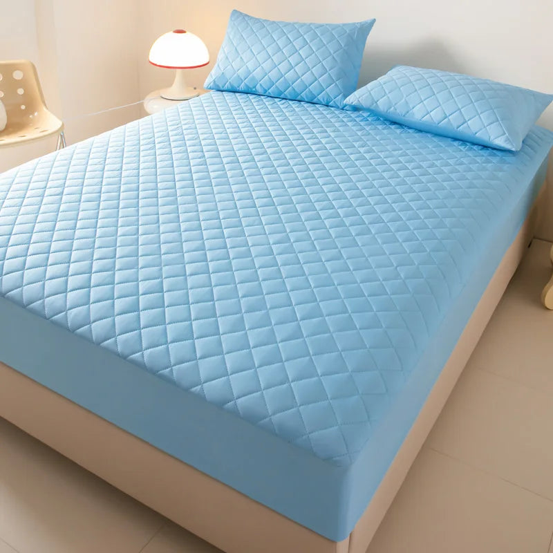 Light blue quilted mattress protector on a bed with matching pillows and a bedside lamp.