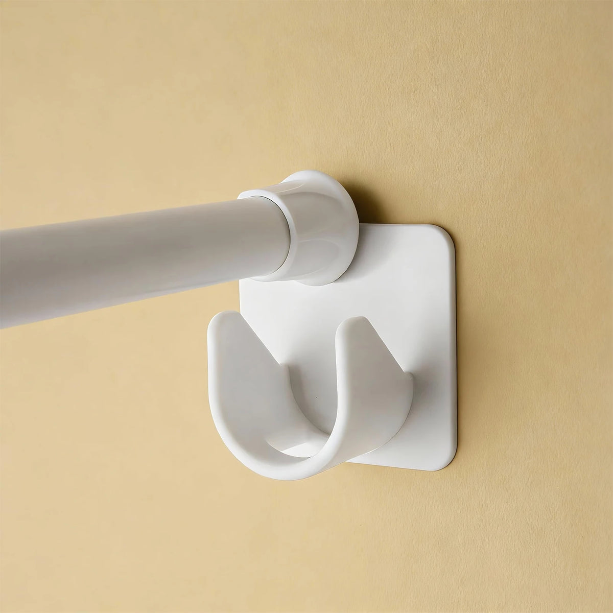 A white self-adhesive curtain rod clip is mounted on a beige wall, with a white rod extending from it.