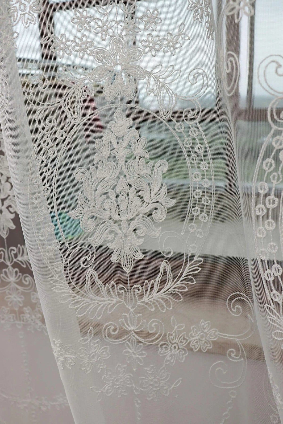 Sheer white embroidered curtains featuring delicate floral and vine patterns in a window setting.