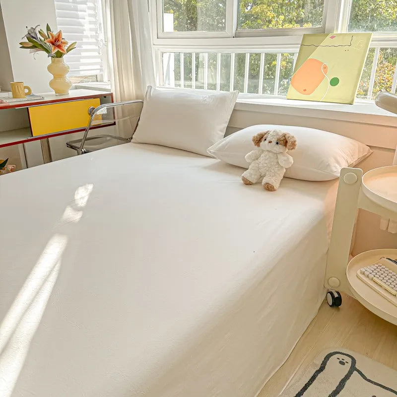 A white organic cotton bedding set with a flat sheet and pillow, featuring a small teddy bear decoration.