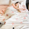 A woman is relaxing on a white quilt with pink floral embroidery, featuring a light pink blanket and pillows.