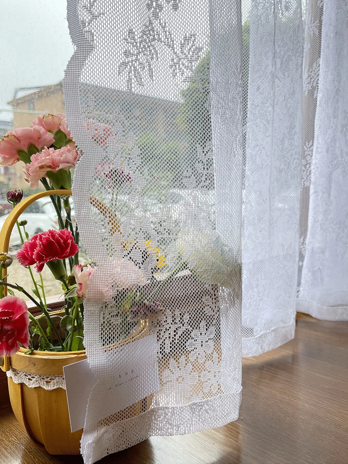 A sheer floral lace curtain panel hangs next to a potted pink flower arrangement.