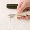 A hand is holding a pair of stainless steel sewing and embroidery scissors next to a spool of dark green thread.
