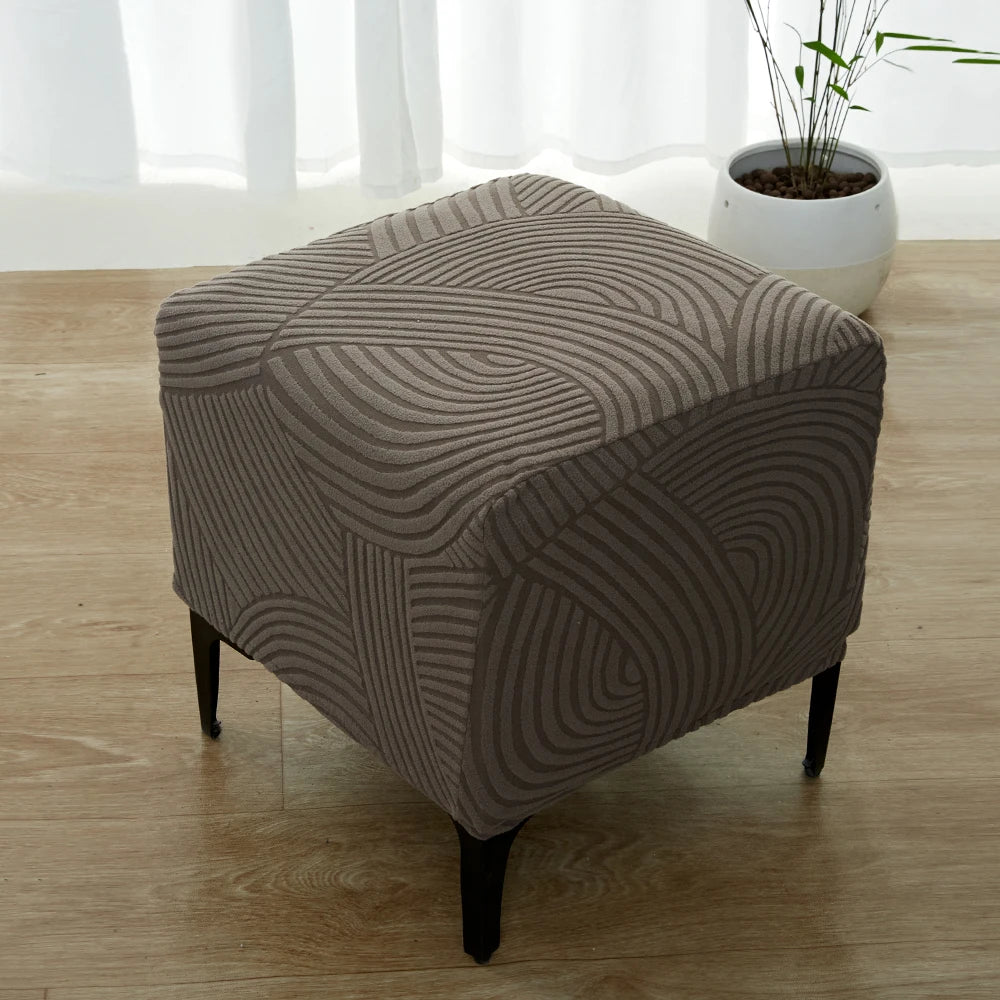 A beige geometric jacquard ottoman slipcover resting on a wooden floor.