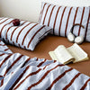 A set of soft washed cotton stripe duvet cover and pillowcases in white and brown stripes with a book and headphones.