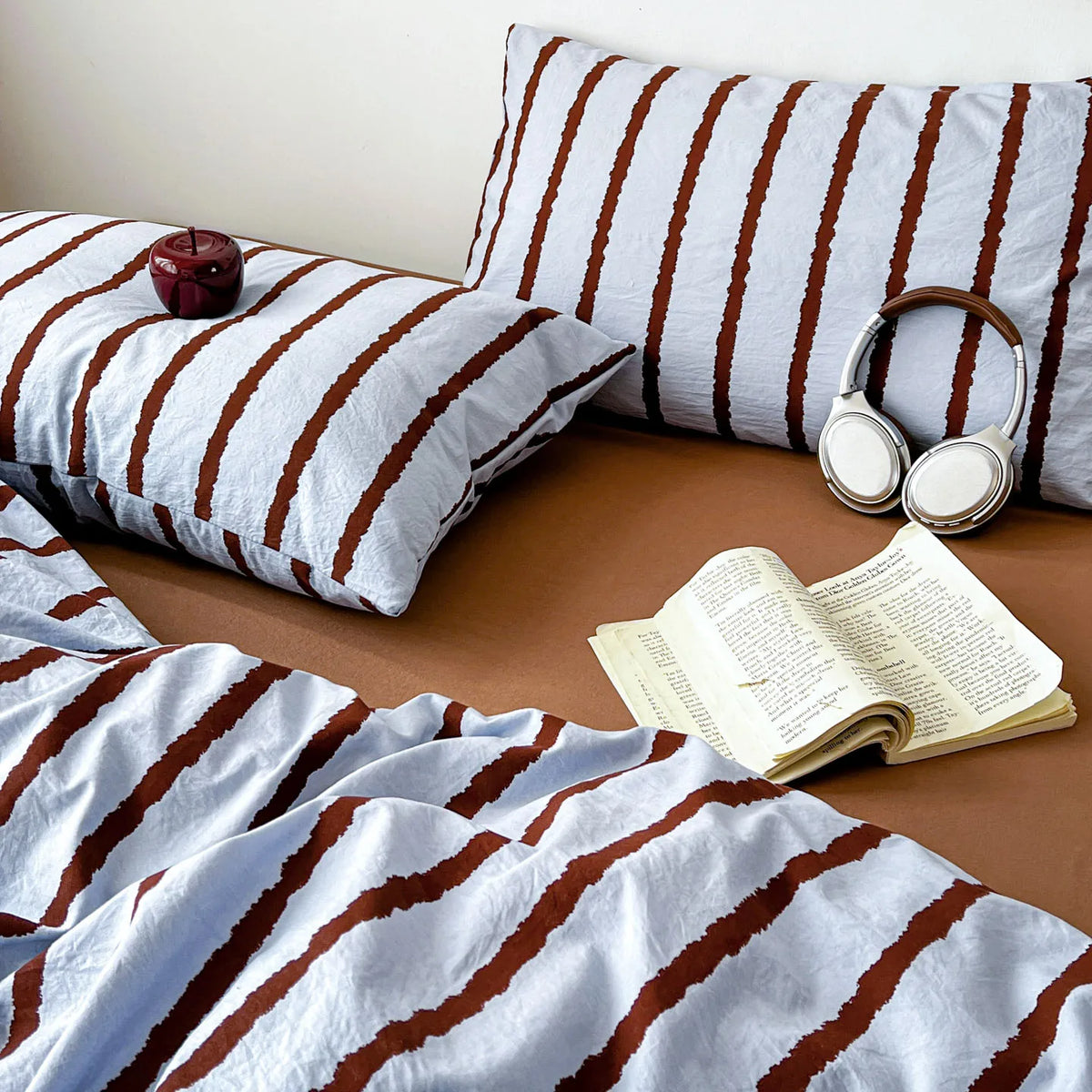 A set of soft washed cotton stripe duvet cover and pillowcases in white and brown stripes with a book and headphones.