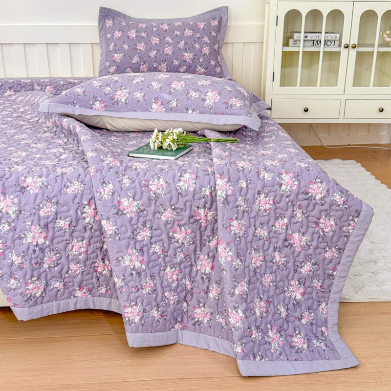 A purple floral quilted bedspread with pink flowers is displayed on a bed, next to a white cabinet.