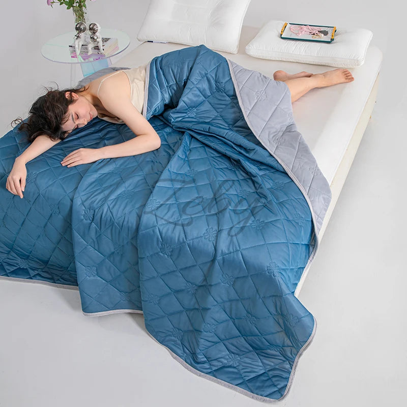 A person is lying on a white surface under a blue quilted blanket, with a small electronic device nearby.