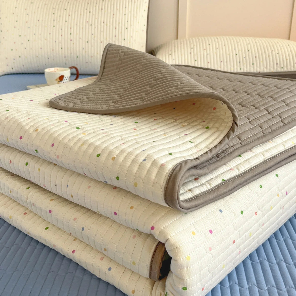 A stack of light beige quilted bedspreads with subtle floral patterns resting on a blue surface.