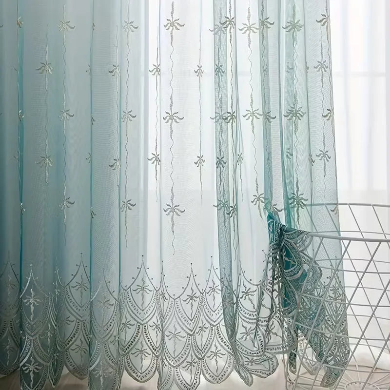 Light blue sheer curtains with silver floral wave embroidery hanging in a window.