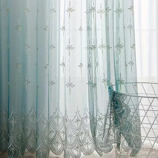 Light blue sheer curtains with silver floral wave embroidery hanging in a window.