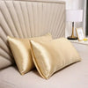 Two golden silk pillow covers resting on a beige headboard with a white lamp.