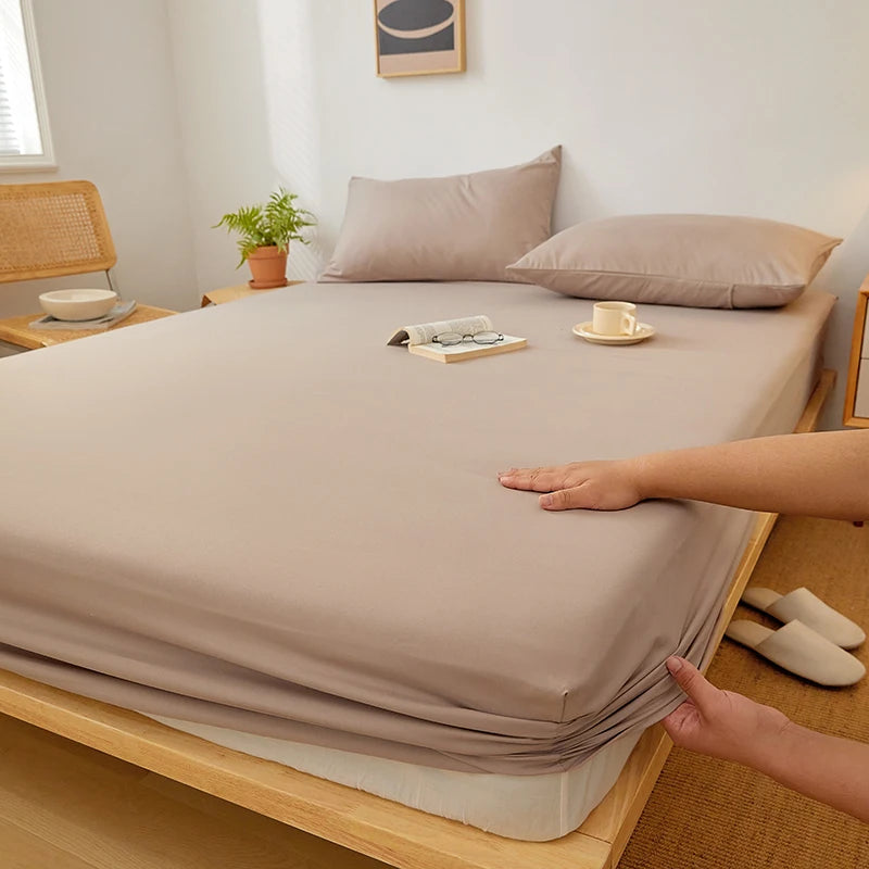 A beige fitted sheet is draped over a wooden bed frame, with a hand adjusting the fabric.