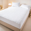 A white quilted mattress protector is displayed on a bed, featuring a diamond pattern.