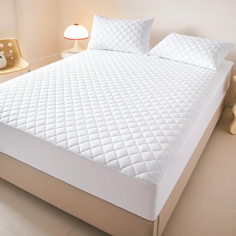 A white quilted mattress protector is displayed on a bed, featuring a diamond pattern.