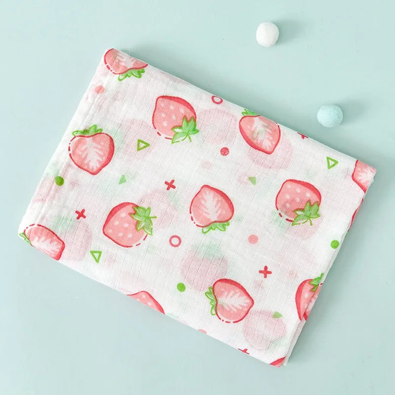 A pink muslin swaddle blanket featuring a pattern of red strawberries and green leaves on a light blue background.