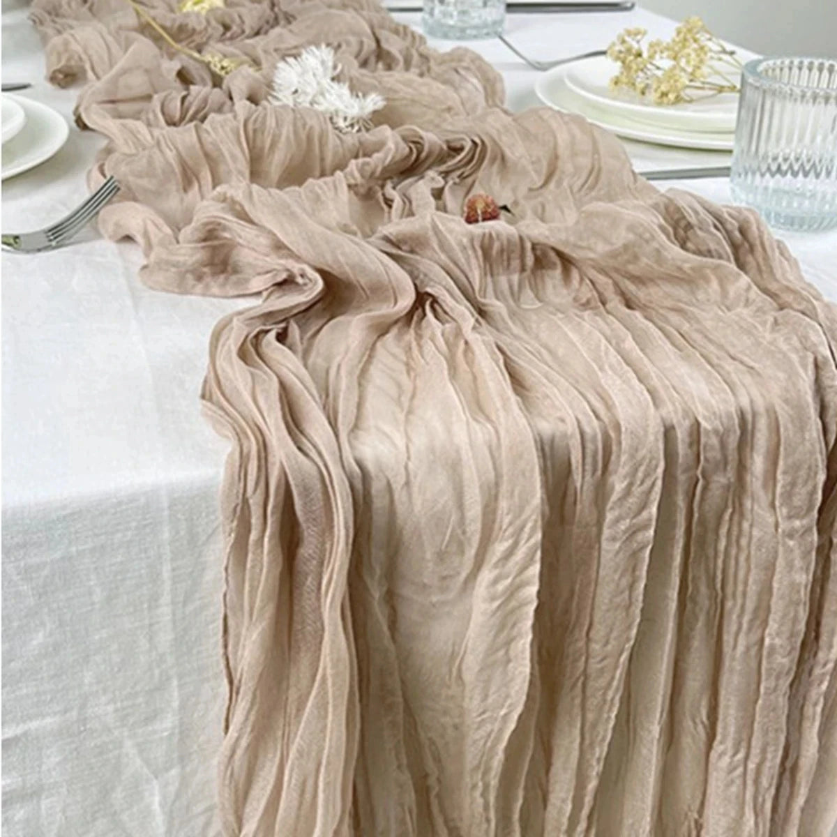 A beige semi-sheer gauze table runner draped elegantly on a white tablecloth with floral accents.