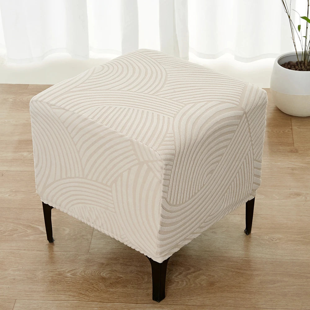 A beige geometric jacquard ottoman slipcover resting on a wooden floor near a window.
