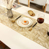 A gold leaf patterned table runner on a white table set with a fried egg, fork, and glass.