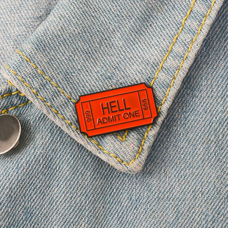 A vintage enamel airline ticket lapel pin with the text HELL ADMIT ONE pinned onto a light blue denim fabric.
