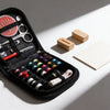 A black sewing kit open to show various hand tools including scissors needles and buttons on a white background.