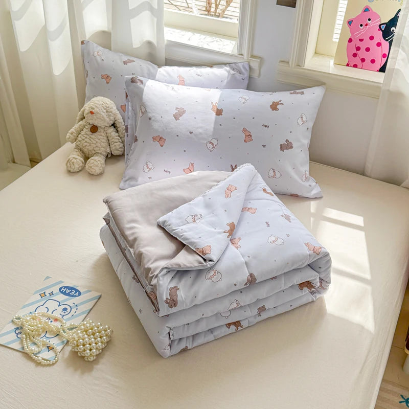 A stack of light purple and white quilted blankets with small animal patterns and a stuffed rabbit are displayed on a table.