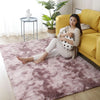 A woman relaxing on a pink fluffy rug in a bright room with large green plants and a yellow sofa.