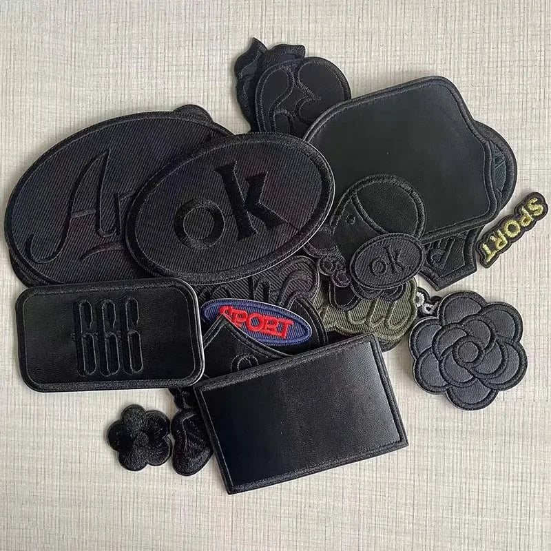 A set of five black embroidered leather patches with self-adhesive backing, including letters, symbols, and floral designs.
