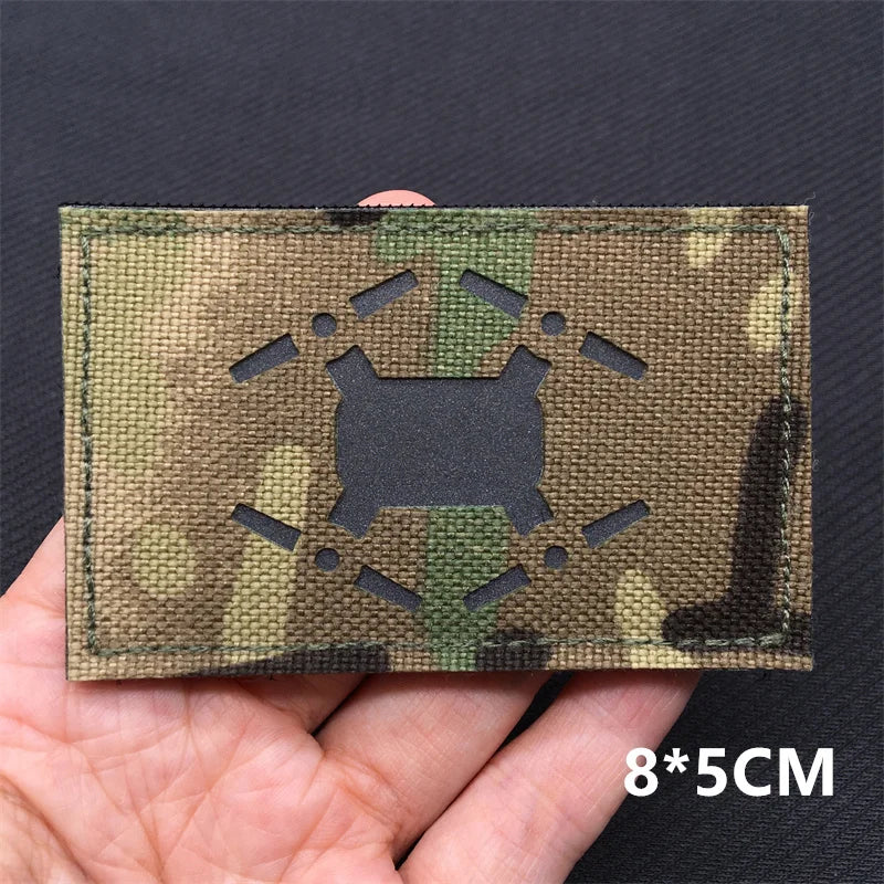 A rectangular tactical patch in a camouflage pattern is held in a hand against a dark background.