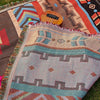 A woven blanket featuring geometric patterns in shades of orange, blue, and beige laid out on grass with a wooden instrument 