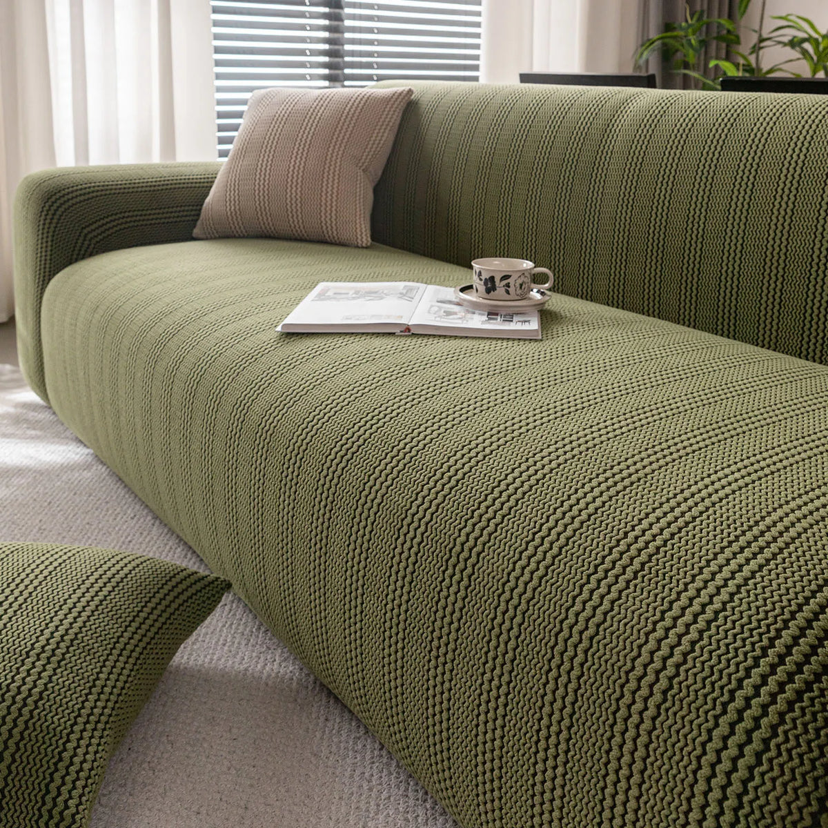 A textured olive green sofa slipcover with a light cushion and a book on it, set in a bright room.