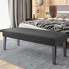 A modern gray velvet bench slipcover with secure straps placed on a light gray striped rug next to a wooden bed.