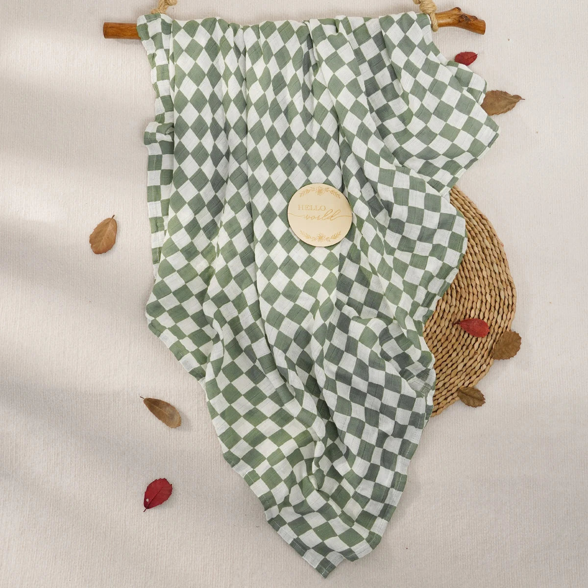 A light green and white checkered muslin swaddle blanket draped over natural wood sticks with dried leaves.