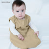 A baby wearing a beige muslin cotton sleeping bag is sitting on a white surface.
